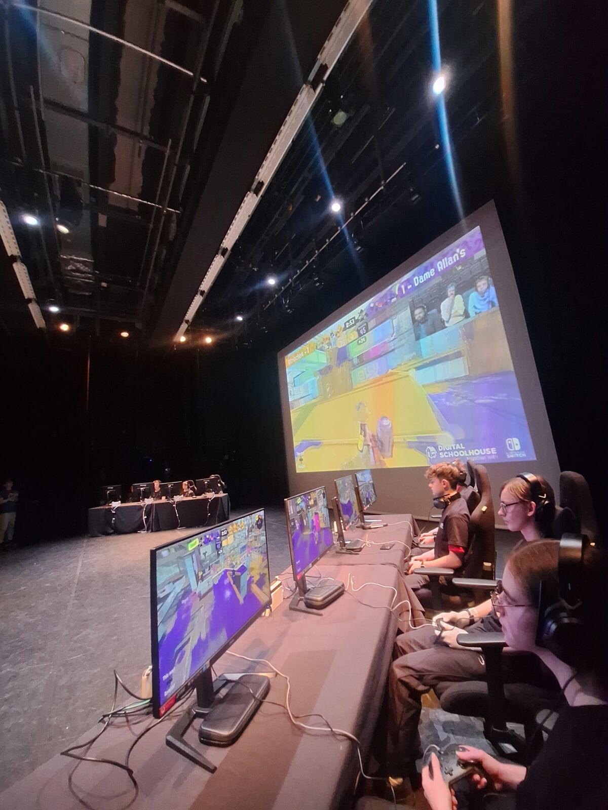 Two teams are locked in competition in a game of Splatoon 3 with a giant screen projecting the game behind them and the stage lights shining down